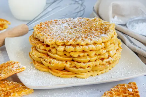 Stapel goldbrauner Waffeln ohne Backpulver, bestäubt mit Puderzucker. Daneben liegen weitere Waffeln.