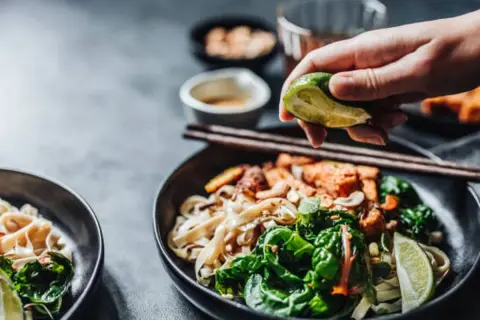 En hand pressar lime över en skål med nudlar och grönsaker, perfekt vin till asiatisk mat.