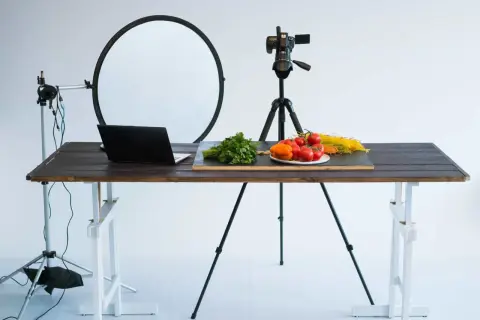 A tabletop photography setup with a camera on a tripod, a laptop, and fresh ingredients like tomatoes and pasta on a wooden table.