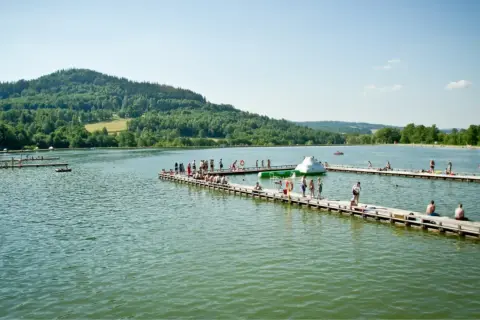 Ludzie odpoczywają na drewnianych pomostach nad jeziorami Dolnego Śląska. W tle zielone wzgórza.