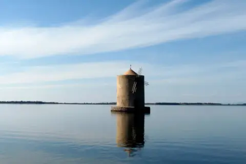 Mulino solitario nella laguna di Orbetello, riflesso sull'acqua calma sotto un cielo azzurro con nuvole sottili.
