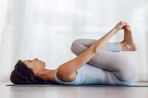 Mujer en postura del bebé feliz, abrazando sus pies con las manos, relajada en una esterilla de yoga.