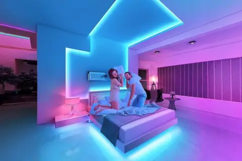 A couple has a pillow fight in a bedroom bathed in vibrant RGB light ideas, with blue and pink hues illuminating the modern space.
