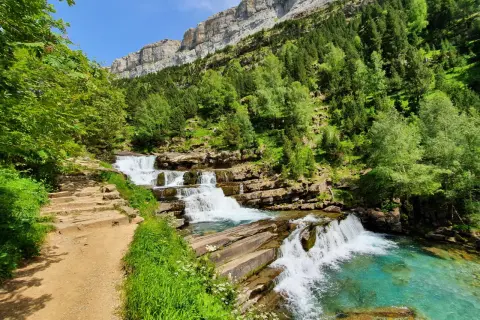 Sendero de piedra junto a las cascadas de las **gradas de soaso**, con vegetación exuberante y un imponente macizo rocoso al fondo.