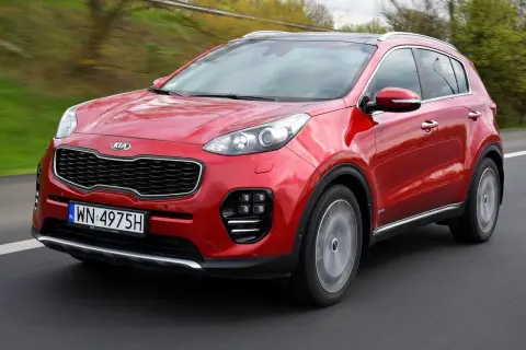 Czerwony Kia Sportage IV pędzi po drodze, otoczony zielenią.