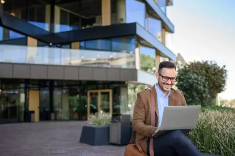 Uśmiechnięty mężczyzna w okularach pracuje na laptopie przed nowoczesnym budynkiem. Może właśnie sprawdza swój dodatek mieszkaniowy.