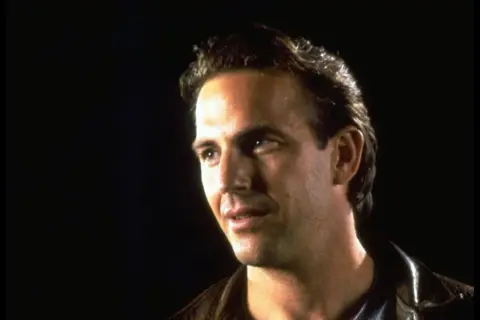 Kevin Costner jung, mit nachdenklichem Blick und brauner Lederjacke vor dunklem Hintergrund.