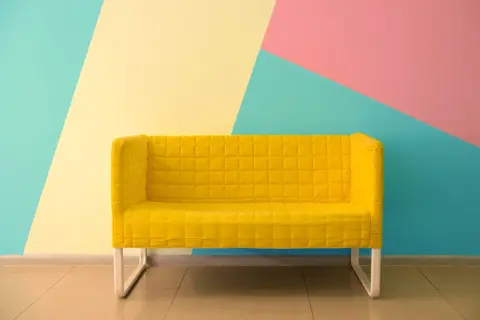 Żółta kanapa na tle geometrycznej ściany w kolorach: miętowym, żółtym i różowym. Świetne inspiracje jak pomalować pokój!