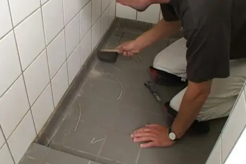 Man knäböjer och använder en borste för att ta bort silikonfog runt en dusch.