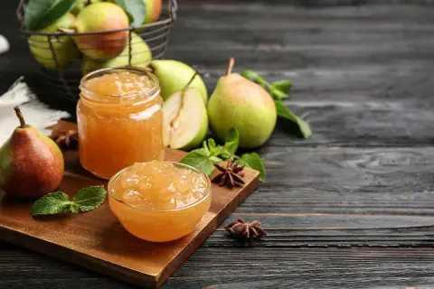 Confiture de poires dans un bocal et un bol, avec des poires fraîches et des feuilles de menthe sur une planche en bois.