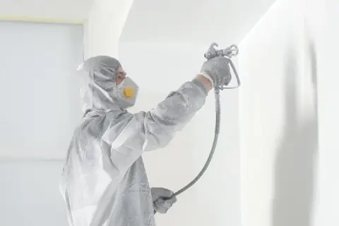 Osoba w kombinezonie i masce ochronnej wykonuje malowanie pistoletem białej ściany.