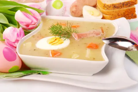 Biały barszcz przepis babci, z jajkiem i kiełbasą, ozdobiony koperkiem. W tle pisanki i tulipany.