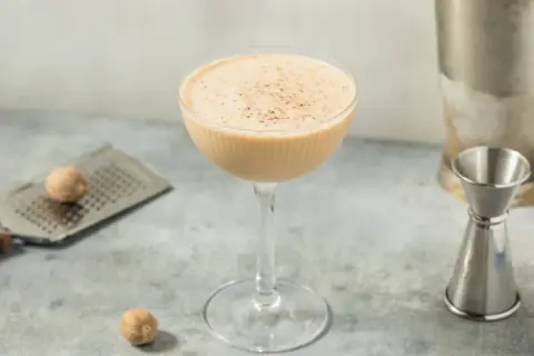 Ein cremiger Brandy Alexander Cocktail in einem Coupette-Glas, garniert mit Muskatnuss, neben einem Schnapsmaß und Muskatnüssen.
