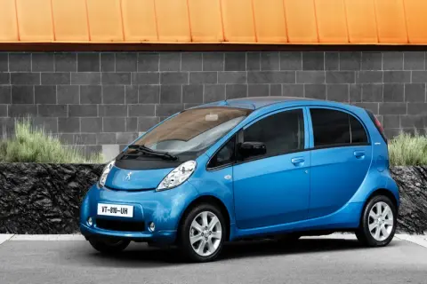 En blå Peugeot iOn står parkerad framför en grå tegelvägg med en orange takkant.