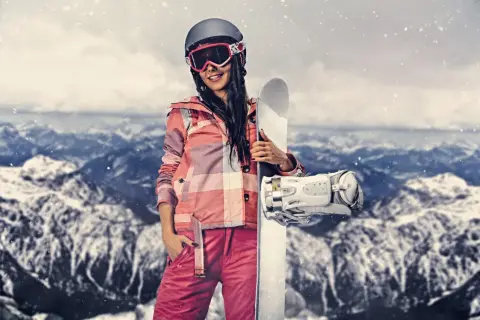 Kobieta w stroju narciarskim trzymająca snowboard na tle gór.