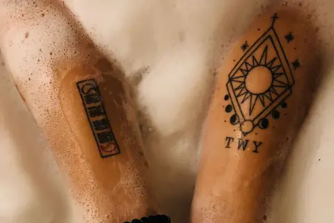 Zwei Arme mit Tattoos in Schaum gebadet. Ein Tattoo zeigt ein geometrisches Muster mit Sonne und Mond, das andere eine Reihe von Symbolen. PH neutrale Seife Tattoo-Pflege.