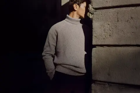 Uomo con un elegante outfit con dolcevita grigio, in piedi vicino a un muro di pietra.