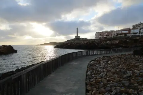 Camino de piedra y madera bordeando el mar, con un faro al fondo. La carretera subida al faro invita a pasear al atardecer.