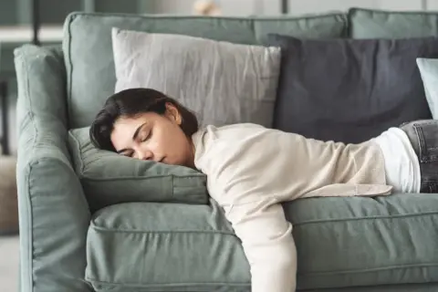 Eine Frau liegt erschöpft auf einem Sofa, ihre Augen sind geschlossen. Sie leidet unter Gliederschmerzen ohne Fieber und ruht sich aus.