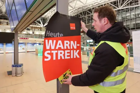 Ein Mann mit Warnweste befestigt ein Schild "Heute WARN-STREIK" an einem Pfosten. Hier wird heute gestreikt.