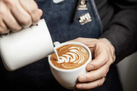 Barista skapar vacker latte art i en vit kopp. Mjölkskummet bildar ett mönster av virvlar.