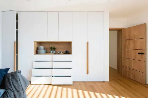Armarios ocultos en pared blancos con nicho de madera y cajones abiertos. Puerta de madera a la derecha.