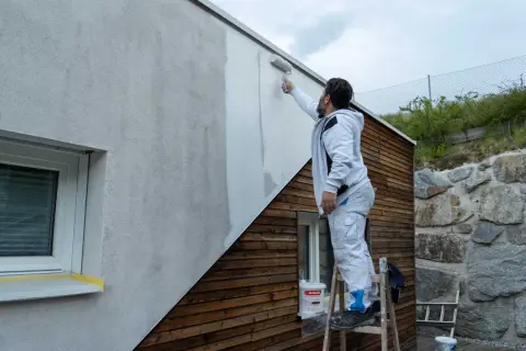 Mann streicht Fassade ohne Reinigung mit Roller. Ein Eimer Farbe steht bereit.