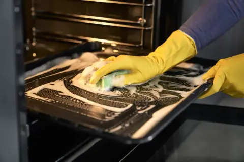 Main tenant avec des gants jaunes, nettoie une plaque de cuisson savonneuse pour enlever odeur four.