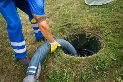 Pracownik w niebieskim kombinezonie wkłada wąż do szamba, co jest częścią procesu jak zrobić odpowietrzenie szamba.