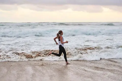 Frau joggt barfuß am Strand entlang, während die Wellen des Meeres an den Sand spülen. Sie genießt ihr tägliches Joggen.
