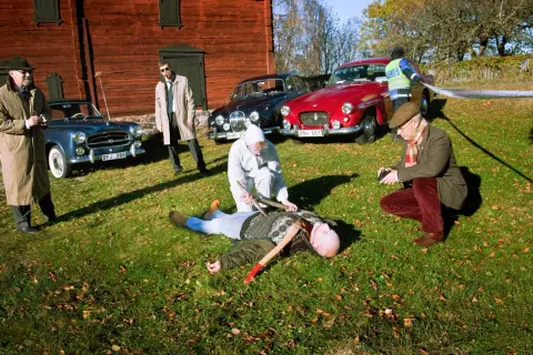 En brottsplats med en död man på marken, omgiven av gamla bilar, inklusive en British Leyland. Polis och utredare arbetar.