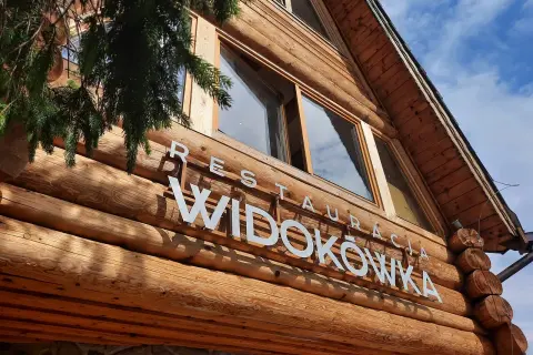 Drewniana restauracja Widokówka z białym napisem. Gałązka świerku zwisa z lewej strony.