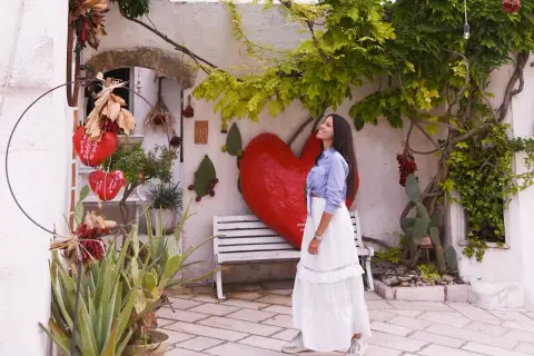 Una donna sorride sotto un albero rigoglioso, con un grande cuore rosso e decorazioni che ricordano il pomodoro ruffano.