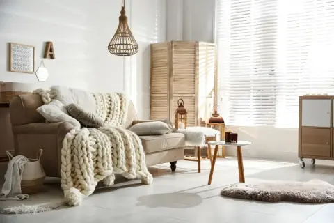 Przytulny salon w stylu hygge wnętrza. Beżowa kanapa z grubym, plecionym kocem, wiklinowa lampa, parawan i świece tworzą ciepłą atmosferę.