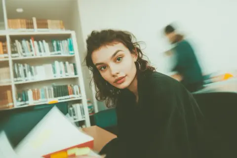 Młoda kobieta w bibliotece, otoczona książkami. Czyta, a obok niej widać ruchliwą postać. Ibiden, co to za miejsce?