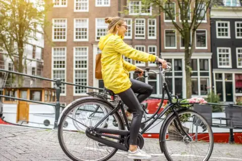 Vrouw in gele jas rijdt op een simpele stadsfiets door Amsterdam, met bloemen in het mandje.