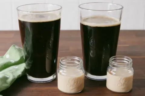 Zwei Gläser dunkles Bier und zwei kleine Gläser mit cremiger Flüssigkeit auf einem Holztisch.