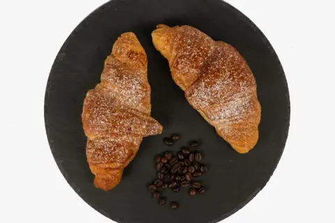 Dos cruasanes dorados espolvoreados con azúcar glas, junto a granos de café. Una delicia para aprender como hacer cruasanes.