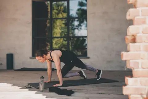Femme en position de planche, genou vers poitrine, effectuant un exercice polyarticulaire. Bouteille d'eau et téléphone à proximité.