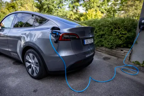Grå elbil laddas med blå kabel från laddbox på väggen. En grönskande häck syns i bakgrunden.