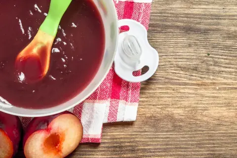 Schüssel mit pflaumenmus baby, Löffel, Schnuller und frische Pflaumen auf Holztisch.