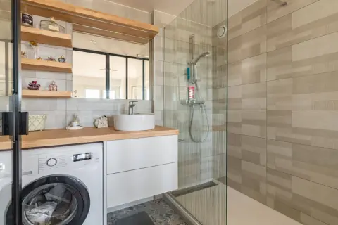 Coin buanderie dans salle de bain moderne avec lave-linge, lavabo sur meuble blanc et douche vitrée.