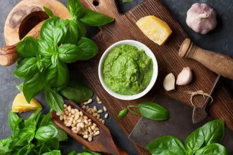 Świeży, zielony sos bazyliowy w miseczce, otoczony listkami bazylii, orzeszkami pinii, czosnkiem i parmezanem.
