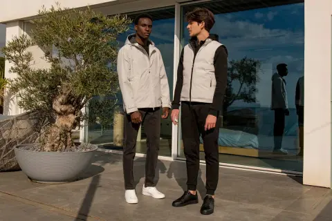 Due uomini con un outfit uomo elegante sportivo, uno con giacca grigia e jeans, l'altro con gilet grigio e pantaloni neri, in piedi all'esterno.