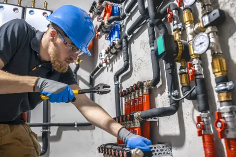 Hydraulik w niebieskim kasku naprawia instalację wodną za pomocą klucza.