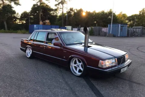 En mörkröd Volvo 760 D24 med en vertikal avgasrör som sticker upp från motorhuven. Bilen har vita fälgar och är parkerad på en asfalterad yta.