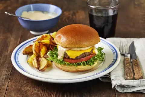 En saftig hamburgare med ost, sallad och tomat, serverad med klyftpotatis och en skål med krämig hamburgerdressing.