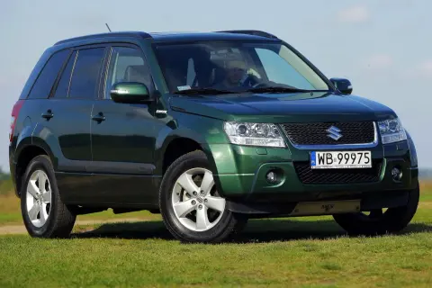 Zielony Suzuki Grand Vitara na tle łąki i błękitnego nieba. Kierowca za kierownicą.