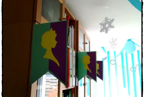 Guirlande "décoration reine des neiges à faire soi même" avec silhouettes jaunes sur fond violet et vert, suspendue devant une bibliothèque.