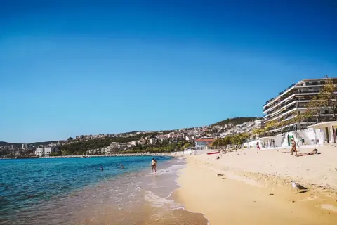 Słoneczna plaża w Grecji, turkusowe morze, białe budynki na wzgórzu. Idealne miejsce na wakacje, gdzie grecja ceny są przystępne.